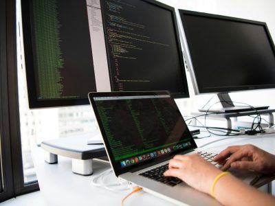 A developer writes code on a laptop in front of multiple monitors in an office setting.